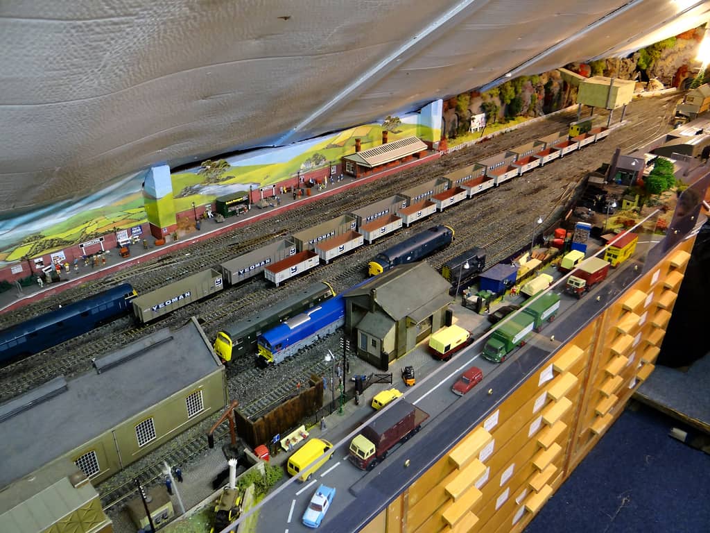 Looking down on a model railway yard: a platform along a painted backscene, rows of grey and white stone wagons, diesels, a shed, a road with lorries, and wooden drawers below.