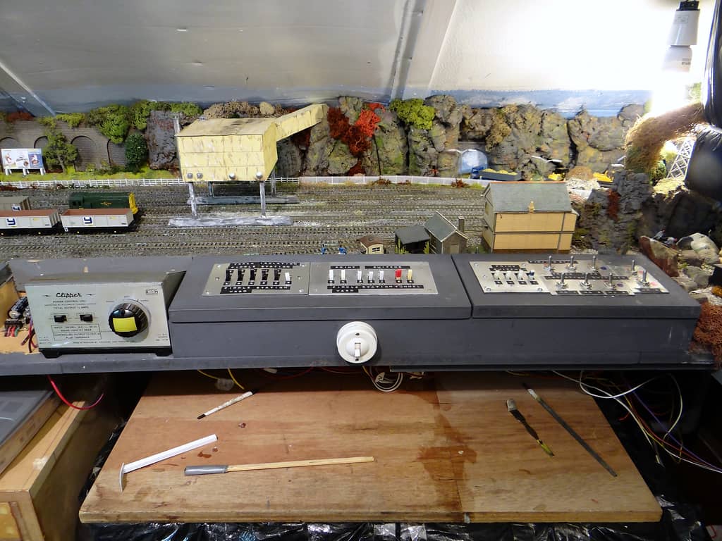 A grey control box with a power unit and rows of switches sits above a wooden bench, with a stone hopper, wagons and rock faces behind.