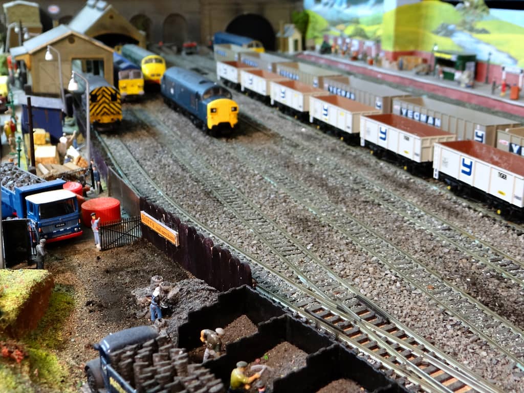 A model coal yard with black timber bunkers, sacks and small figures in front, a blue tipper lorry by an iron gate, and sidings with diesels and white wagons beyond.