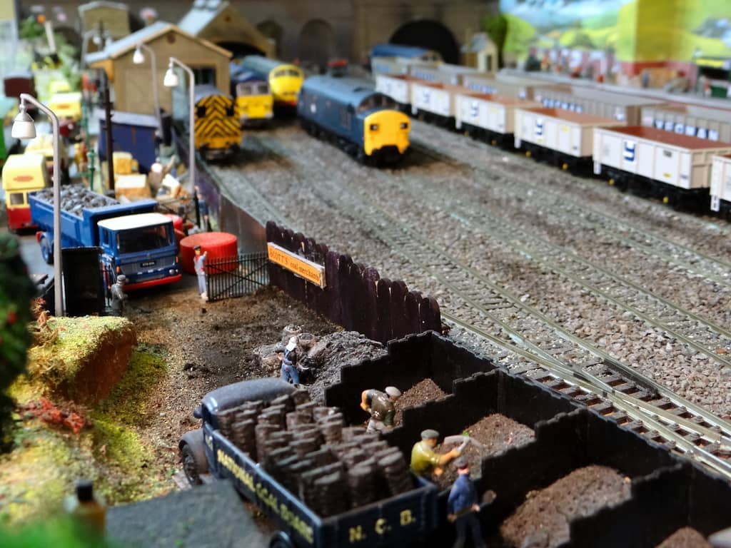 Close view of model coalmen with a pick and shovels in black timber bunkers, beside a dark blue lorry lettered National Coal Board and loaded with sacks of coal.