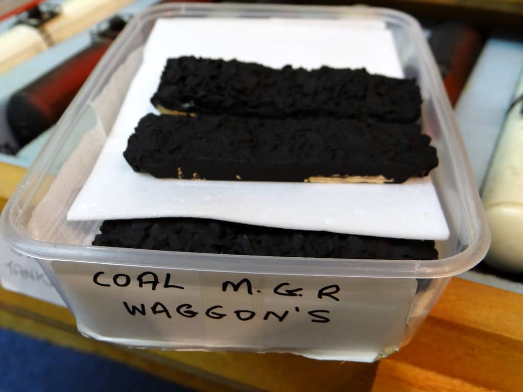 A clear plastic tub labelled COAL M.G.R WAGGON’S, holding long strips of black coal load laid on white sheets.