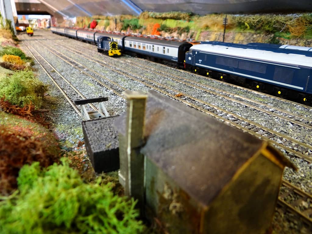 Ground-level view along model sidings: a blurred hut roof in front, a long blue diesel on the right, a small shunter and blue and grey coaches, and a bridge far off.