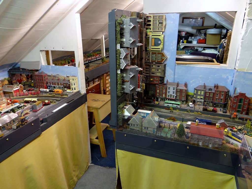 A strip of model town baseboard stands upright on its end, its buildings turned sideways, beside a gap looking through to an aisle with drawers and a stool.