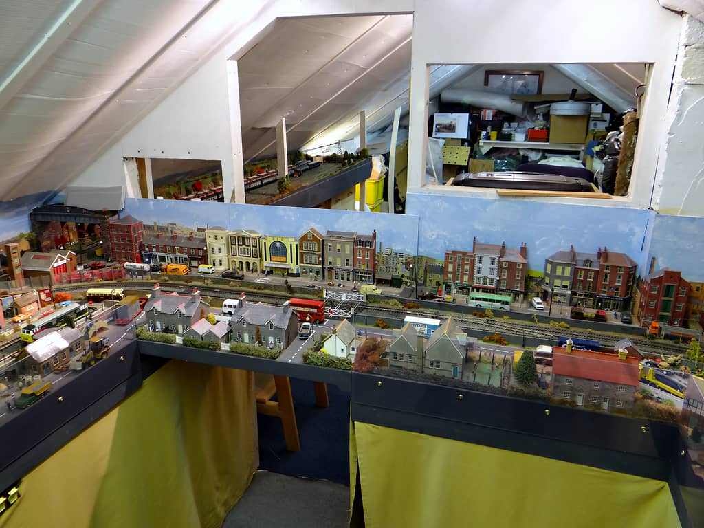 A long model high street with shops, a cinema and a level crossing runs beneath a sloping loft roof, with yellow curtains below and openings in the wall above.