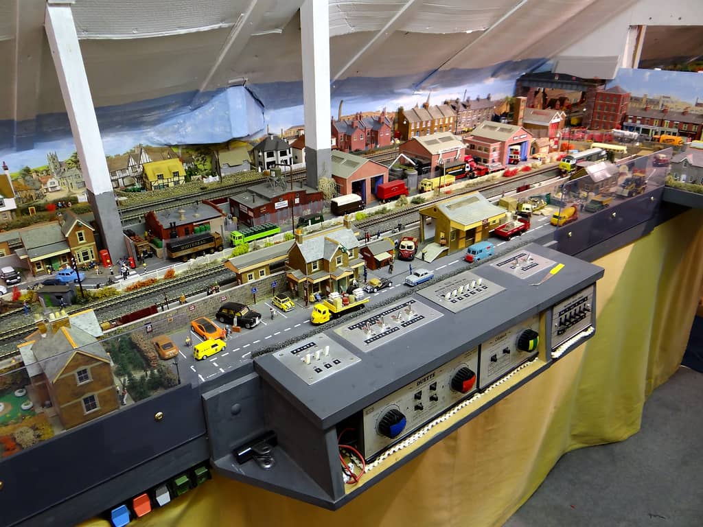 A grey control panel with two power units and switch plates, in front of a model street with parked cars, lorries, a black taxi and a yellow three-wheeled lorry.