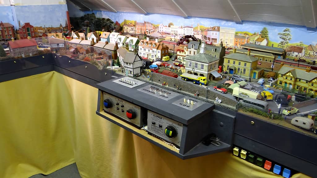 A grey control panel with two power units, their knobs taped blue, red, and green and yellow, in front of a model market town street.