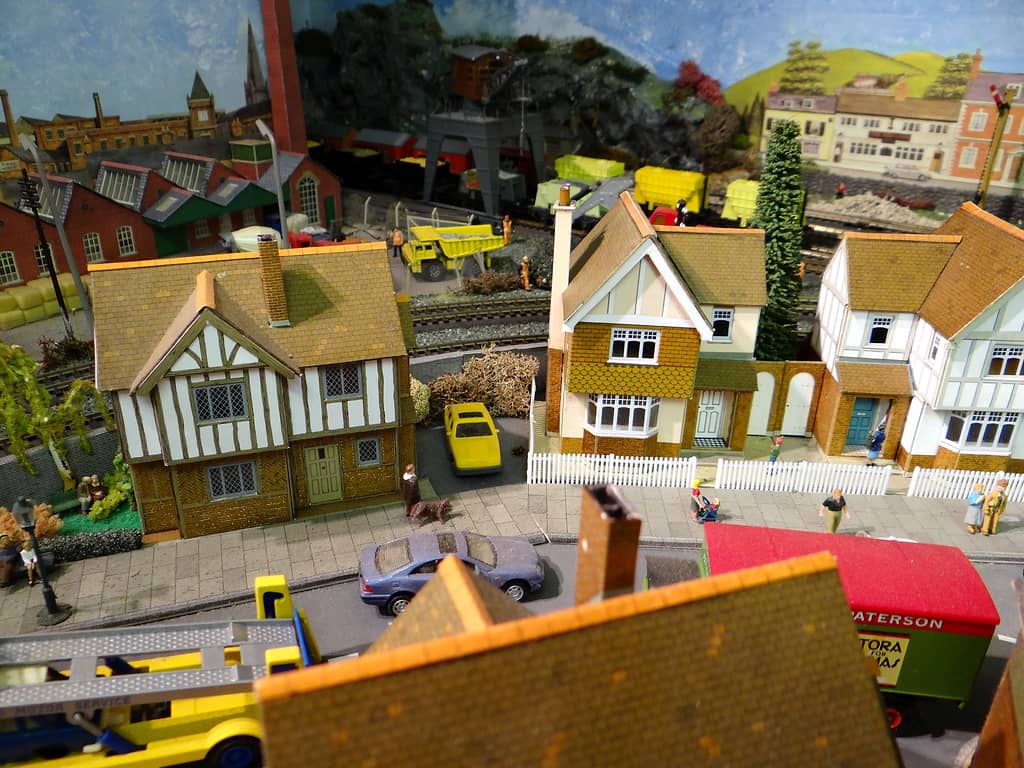 Model street of mock-Tudor and tile-hung houses with white picket fences and people on the pavement, and a factory, chimney and gantry crane beyond the railway.