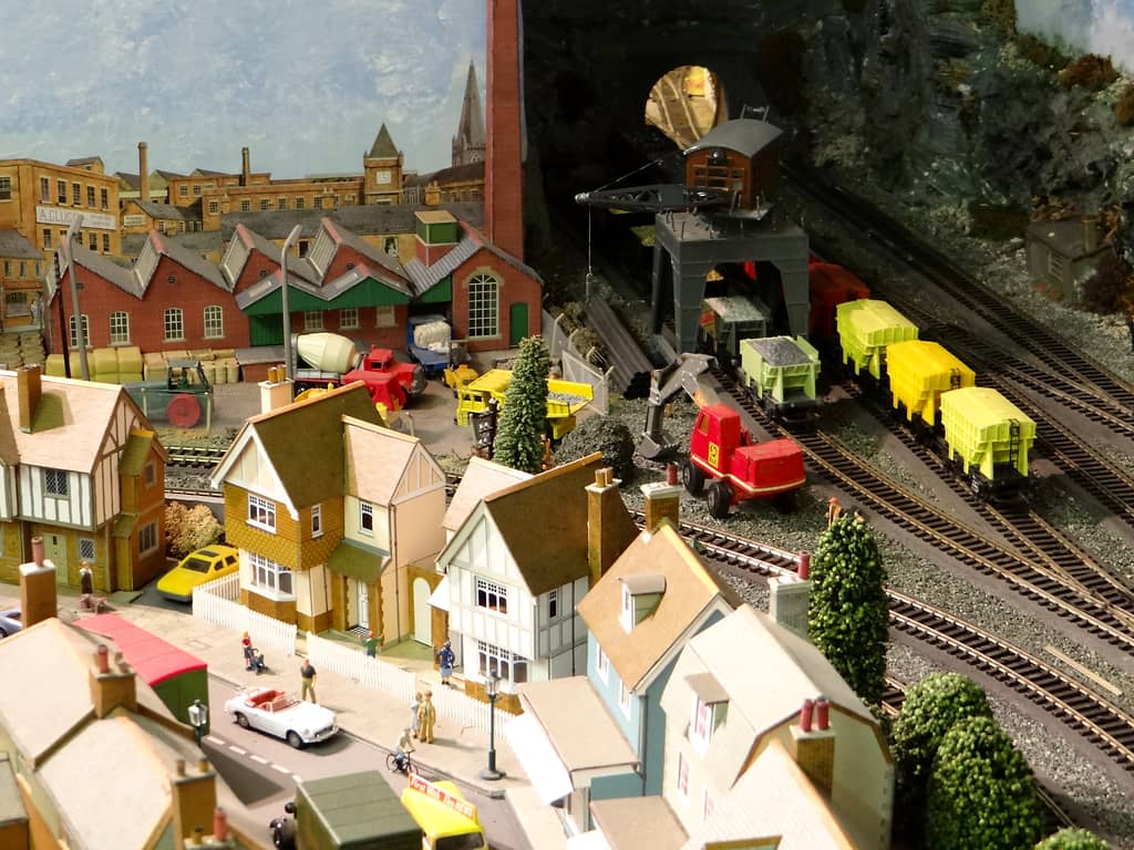 A model street of mock-Tudor houses in front of a red-brick factory with a sawtooth roof, and a grey gantry crane over yellow wagons beside a dark tunnel.