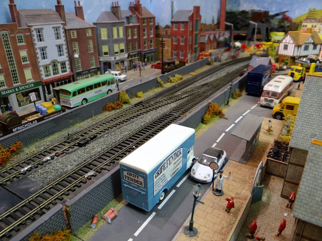 Model street beside a double railway line, with a pale blue box van and a white sports car stopped by a lollipop man, and shops across the tracks.