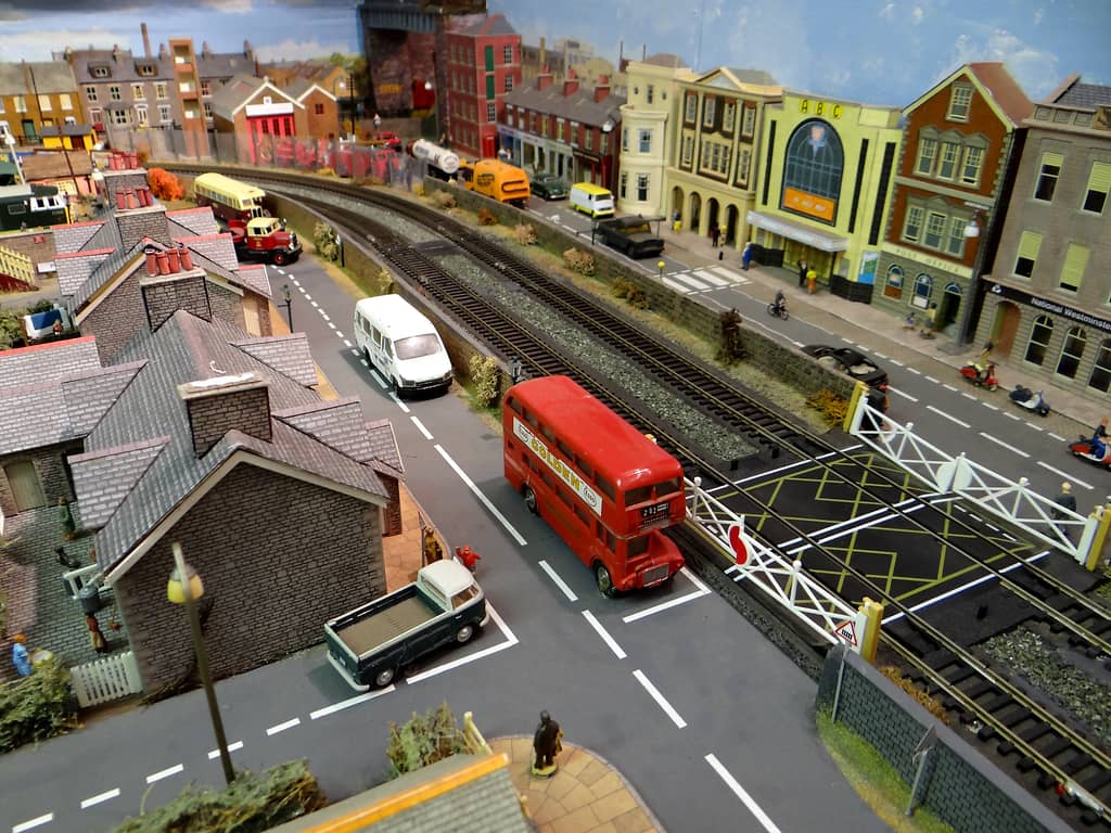 Model level crossing seen from above, with a red double-decker bus near the white gates, stone cottages on the left and a cinema and shops beyond the tracks.