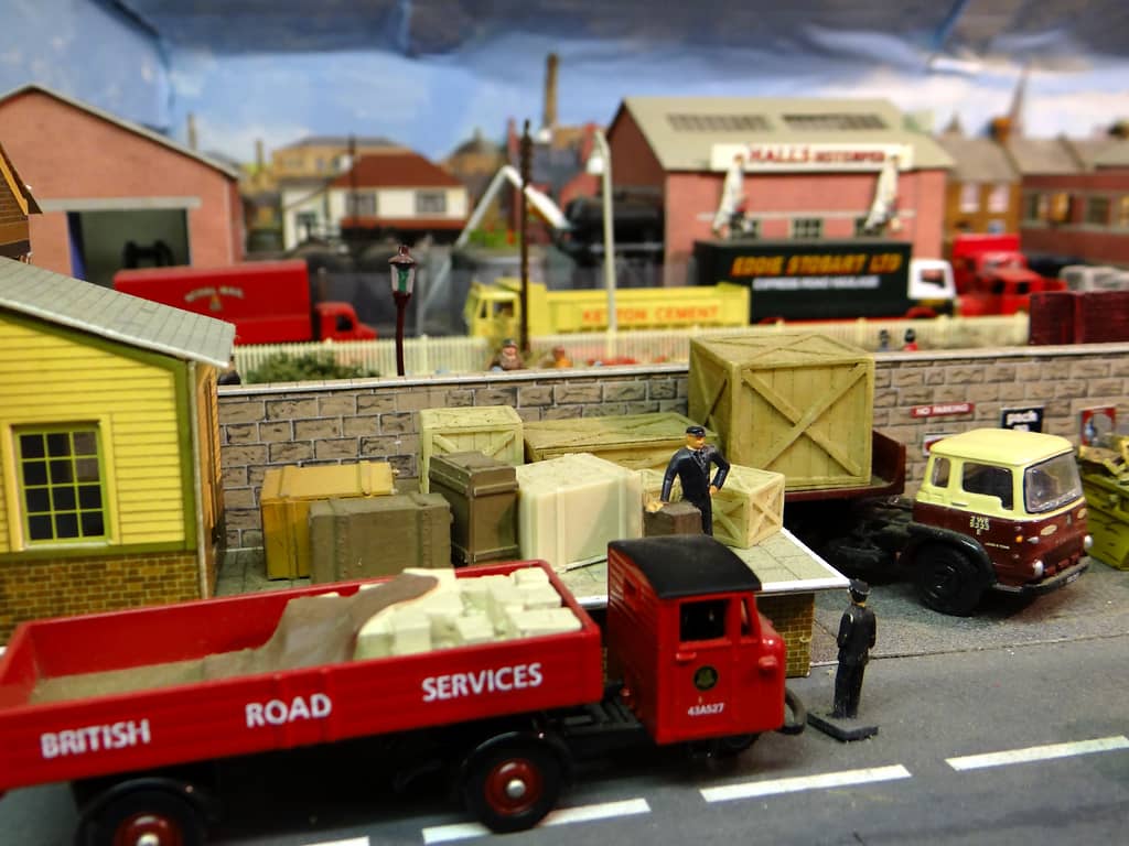 A red British Road Services lorry beside a goods platform stacked with wooden crates, a man in uniform among them, with more lorries parked beyond a stone wall.