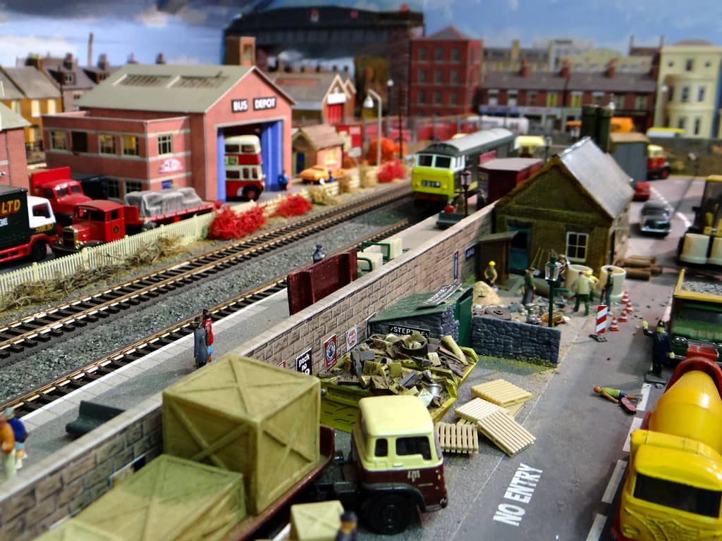 A long view beside a railway line: a stone wall hung with enamel signs, a scrap yard, roadworks with cones, parked lorries and a bus depot in the distance.