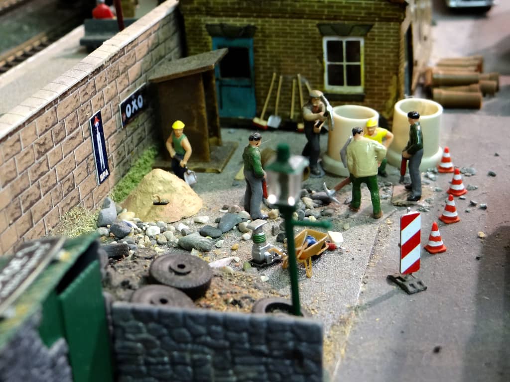 Model workmen with pickaxes, drills and shovels digging beside a stone railway wall, among concrete pipes, a sand heap, a wheelbarrow and traffic cones.