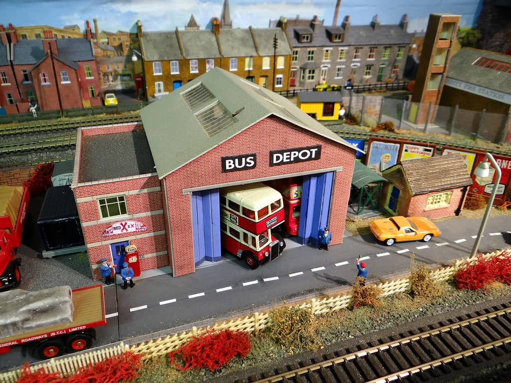 A red-brick model bus depot signed “BUS DEPOT”, its blue doors open on two red double-deckers, with uniformed busmen outside and terraced houses behind.