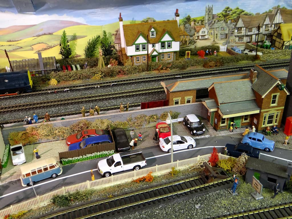 A model red-brick station with a canopy beside a road of parked cars, a VW camper van and a white pickup, with railway tracks and a large white house behind.