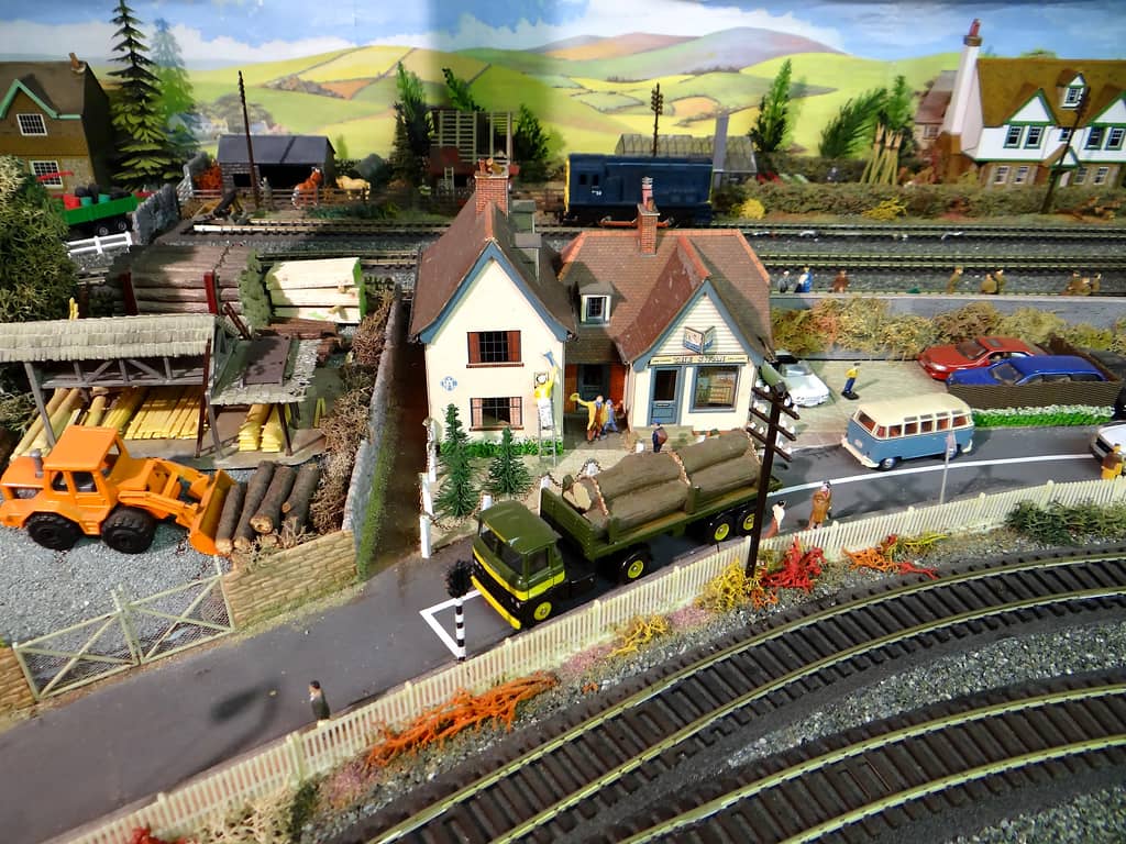 A green lorry loaded with logs outside a white pub, The Swan, beside a timber yard with an orange wheeled loader, planks and log piles, and a road with a VW camper beyond.