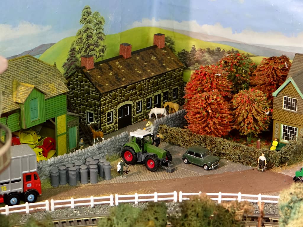 A dark stone farmhouse with a green timber barn, horses behind a stone wall, a green tractor with red wheels, a group of grey milk churns and a green Mini by a hedge.