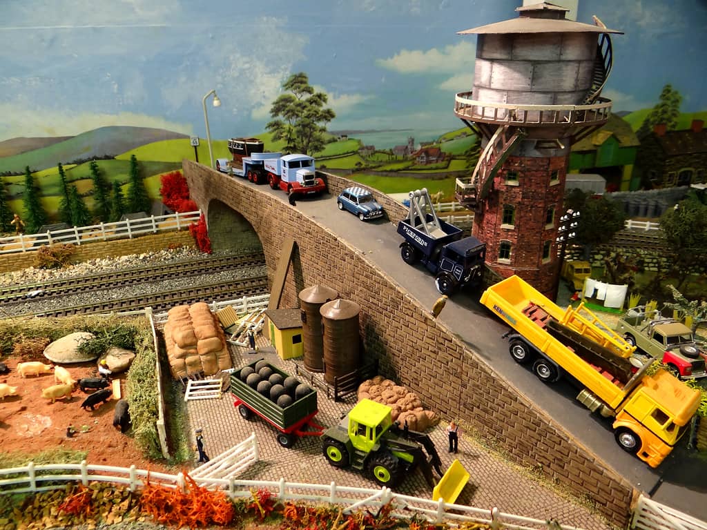 A stone road bridge over railway tracks with lorries and a Mini on it, beside a tall brick water tower, above a pig field, silos, sacks and a tractor and trailer.