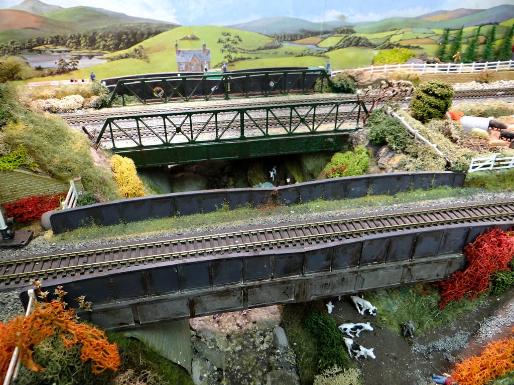 Layered model railway bridges: a grey girder bridge carrying one track in front, green lattice girder bridges behind, and black and white cows beside a stream below.