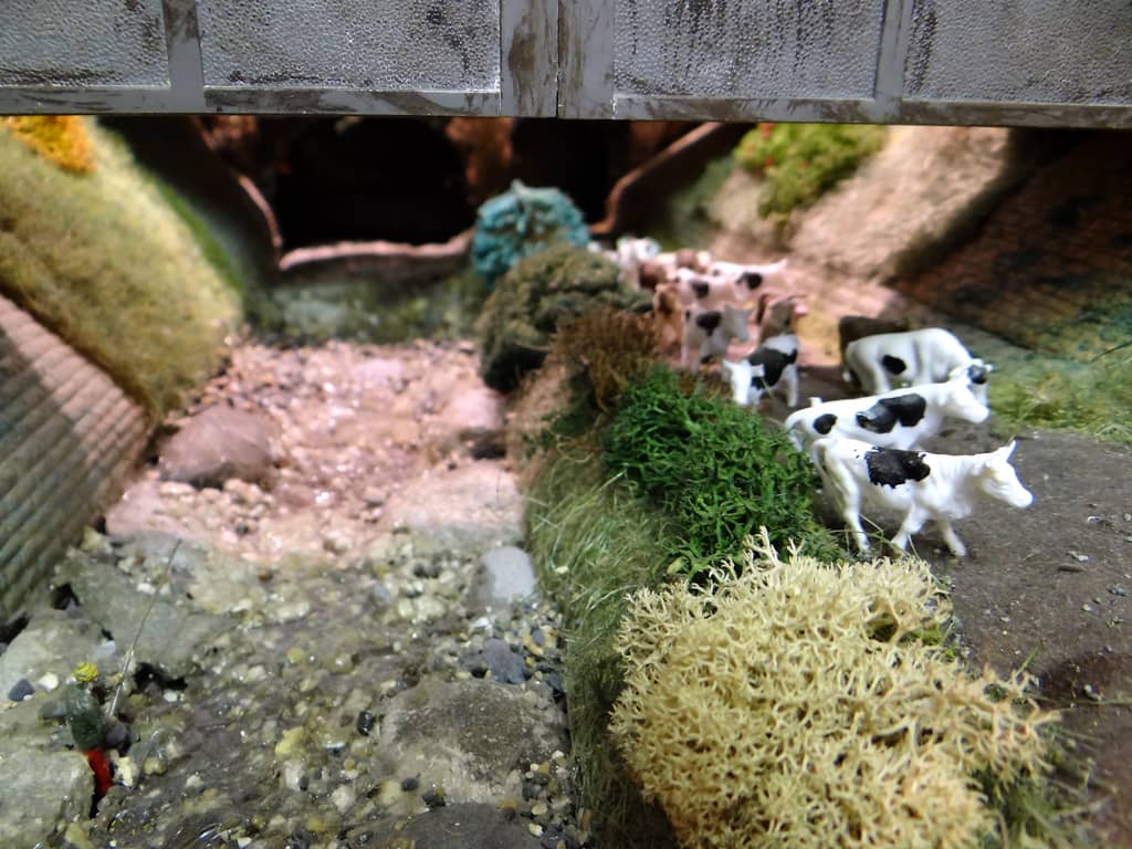 A low close-up of a model stream bed of pale gravel and boulders under a bridge, with black and white cows standing on the bank to the right.