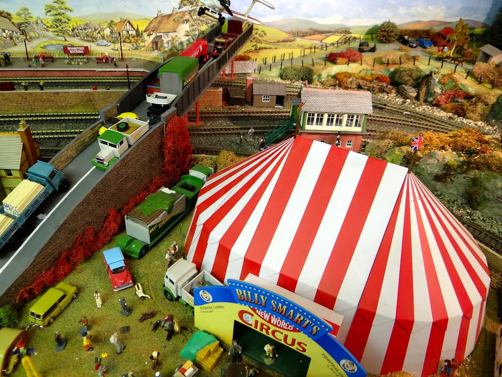 A red and white striped big top seen from above, with a yellow entrance arch, a crowd on the grass, green circus lorries, and a road bridge and railway platform beyond.