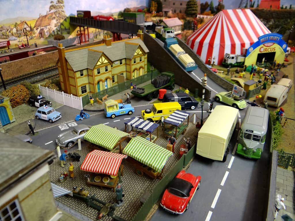 Striped market stalls beside a road with a cream lorry, a grey and green coach and a red sports car, yellow-brick houses behind, and a red and white big top in the far corner.