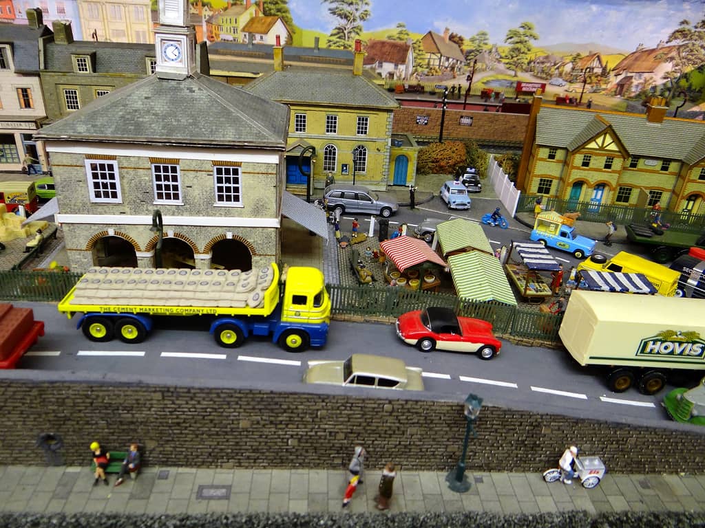 A yellow and blue flatbed lorry loaded with cement sacks outside a stone market hall with a clock tower, with market stalls, police cars and houses behind and a stone wall in front.