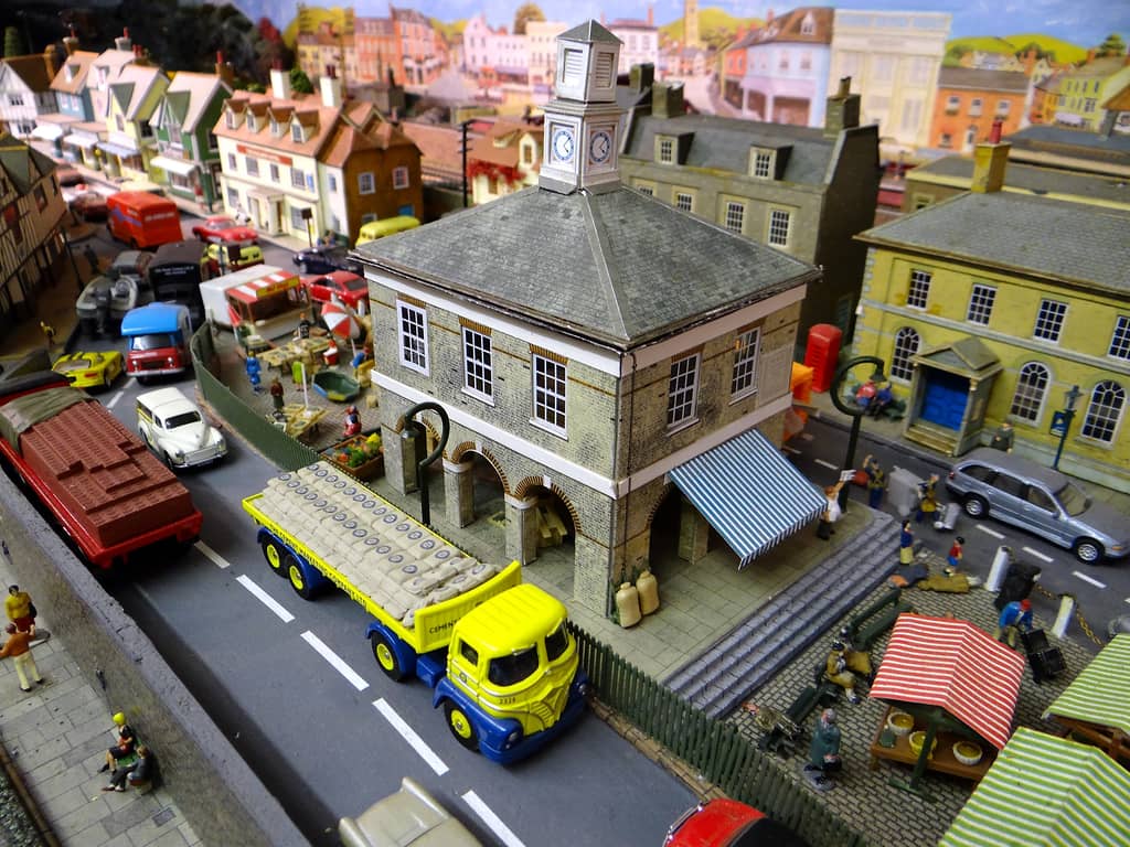 A stone market hall with a white clock tower seen from above, a yellow and blue lorry of cement sacks beside it, a lorry of bricks, market stalls and a street of shops.