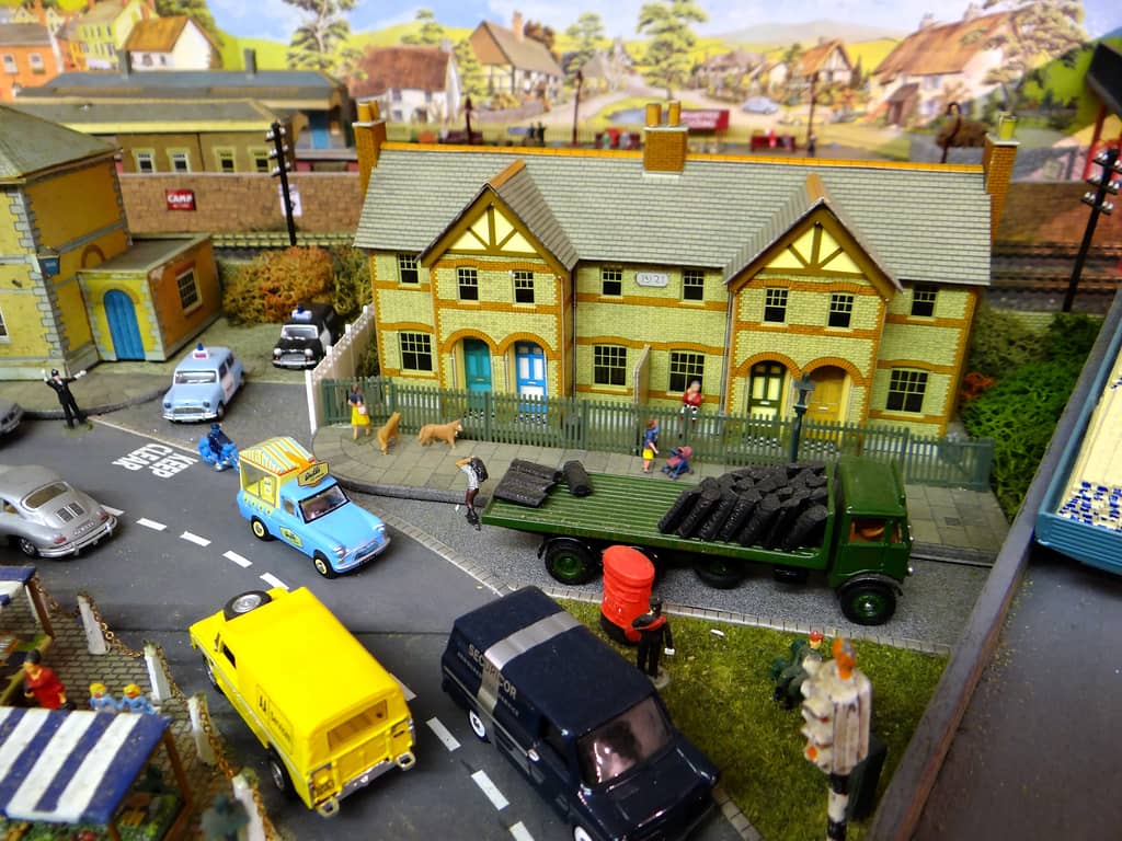 A green flatbed lorry loaded with coal sacks outside a row of yellow-brick houses, with a red pillar box, a navy Securicor van and a yellow Land Rover in the foreground.