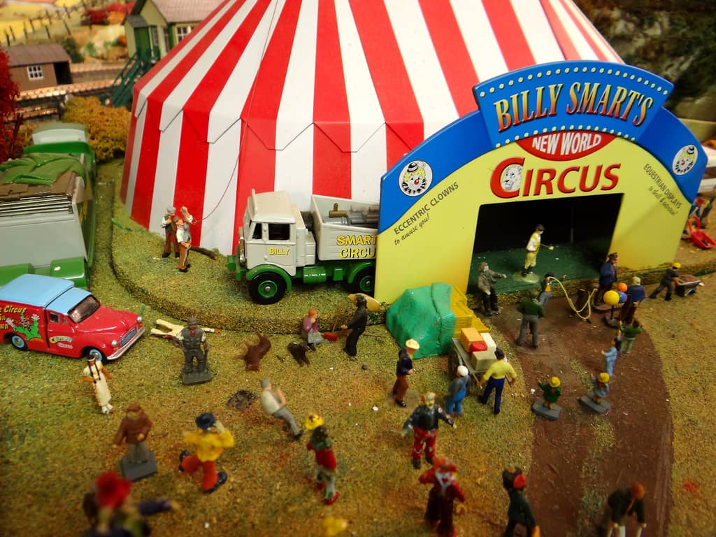 A yellow and blue circus entrance arch lettered BILLY SMART’S NEW WORLD CIRCUS in front of a red and white big top, with a crowd, clowns and circus vehicles on the grass.