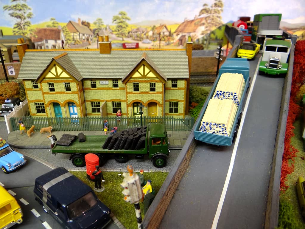 A blue lorry loaded with timber on a ramp beside a row of yellow-brick houses, with a green coal lorry, a red pillar box and a navy van in the street below.