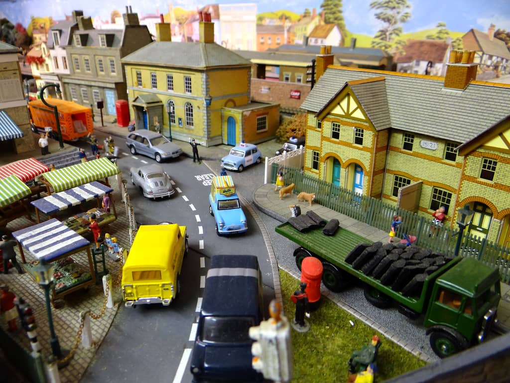 A street of model vehicles running up to a yellow-brick police station, with striped market stalls on the left and a green coal lorry and houses on the right.