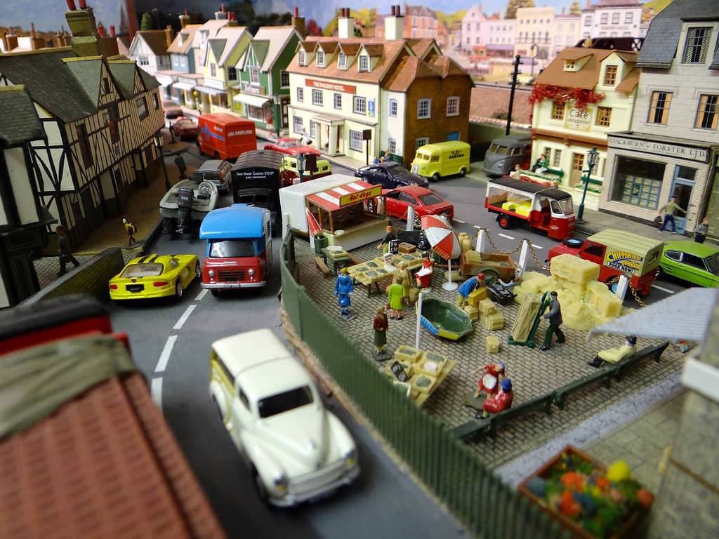 Model market square with crates, stalls, traders and a hot-dog stand, cars and vans parked around it, and a hotel and shops behind.