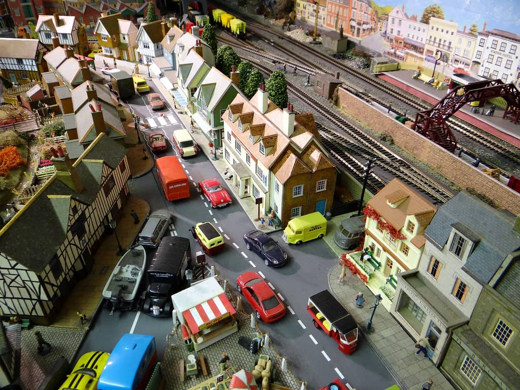 Model town from high above: a busy road of cars and vans between a market stall and a café, a cream hotel, and railway tracks with a red footbridge beyond.