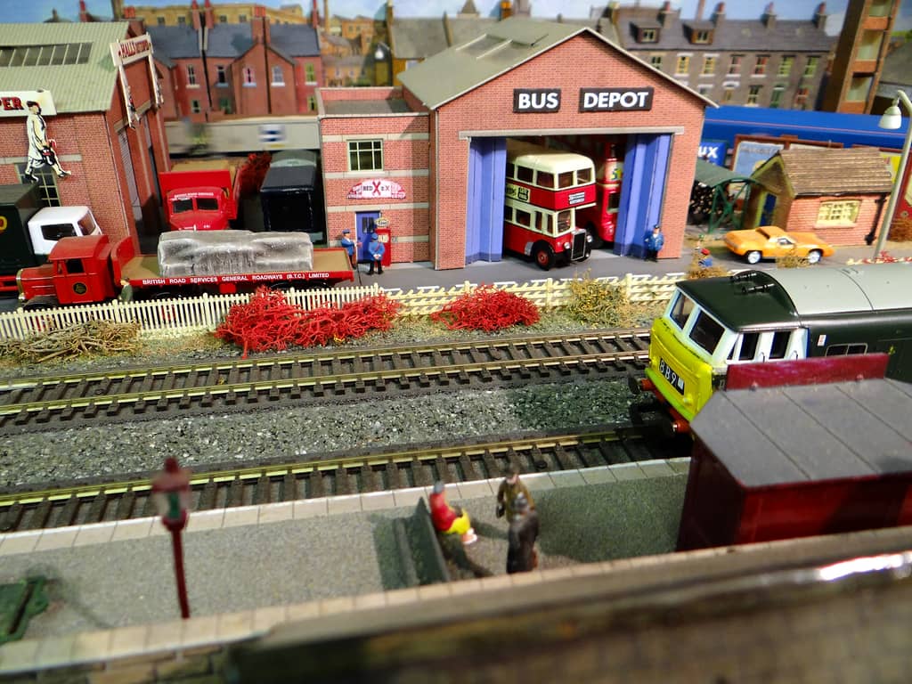 A red-brick model bus depot with red double-deckers, seen across two tracks from a station platform, with red lorries beside it and a diesel’s yellow nose at right.