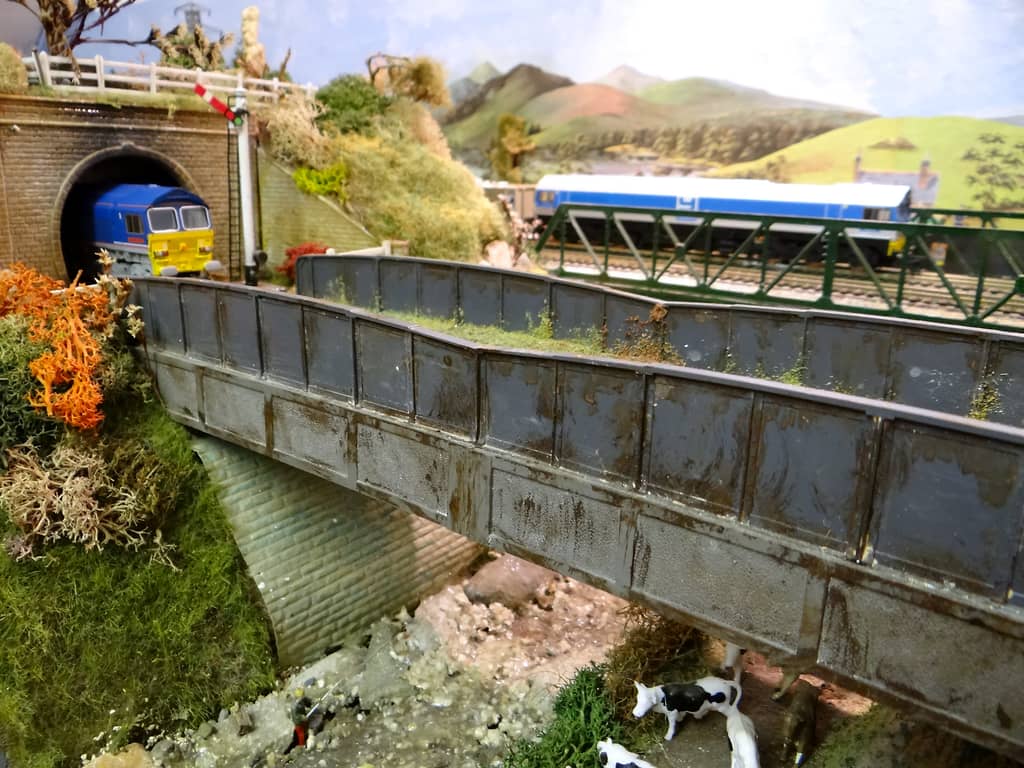 A blue diesel with a yellow front emerges from a tunnel above a weathered grey girder bridge over a rocky stream, with cows below and a blue train on a green lattice bridge behind.