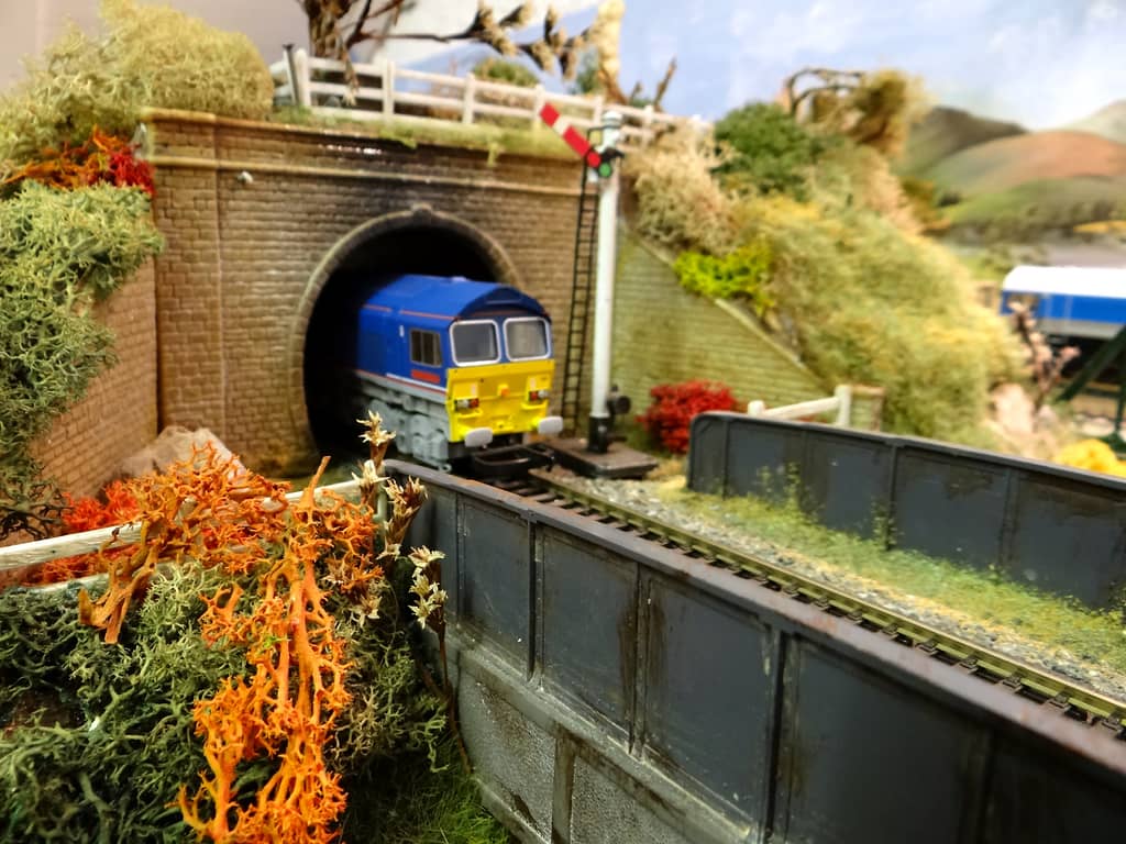 A bright blue diesel locomotive with a yellow front emerges from a tunnel mouth beside a semaphore signal, with orange lichen and a grey plate girder bridge in the foreground.