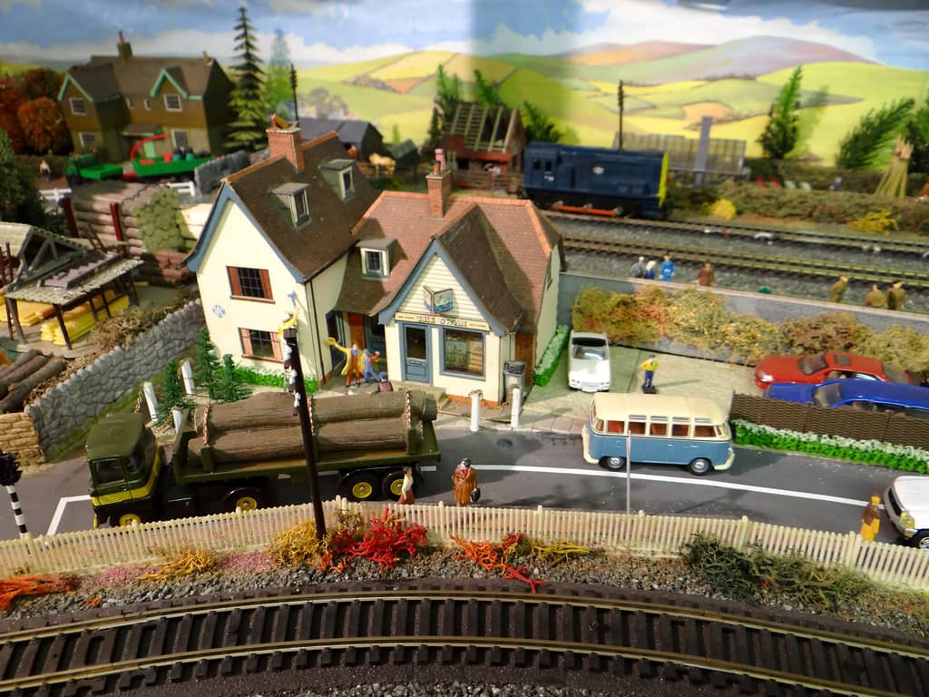 A white pub with a swan sign stands between cottages on a road where a blue-and-cream VW camper and a green log lorry are parked, with railway tracks and a blue shunter behind.