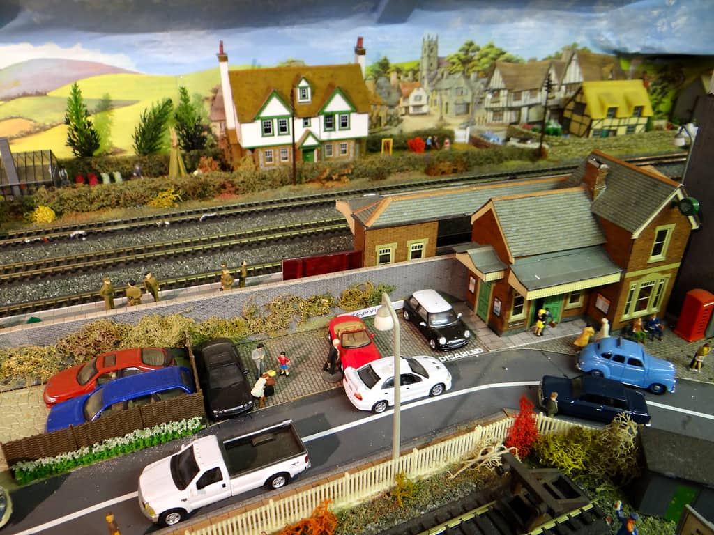 A red-brick station with a fringed canopy and green doors beside a cobbled car park full of model cars and figures, with soldiers on a platform behind a wall and tracks beyond.