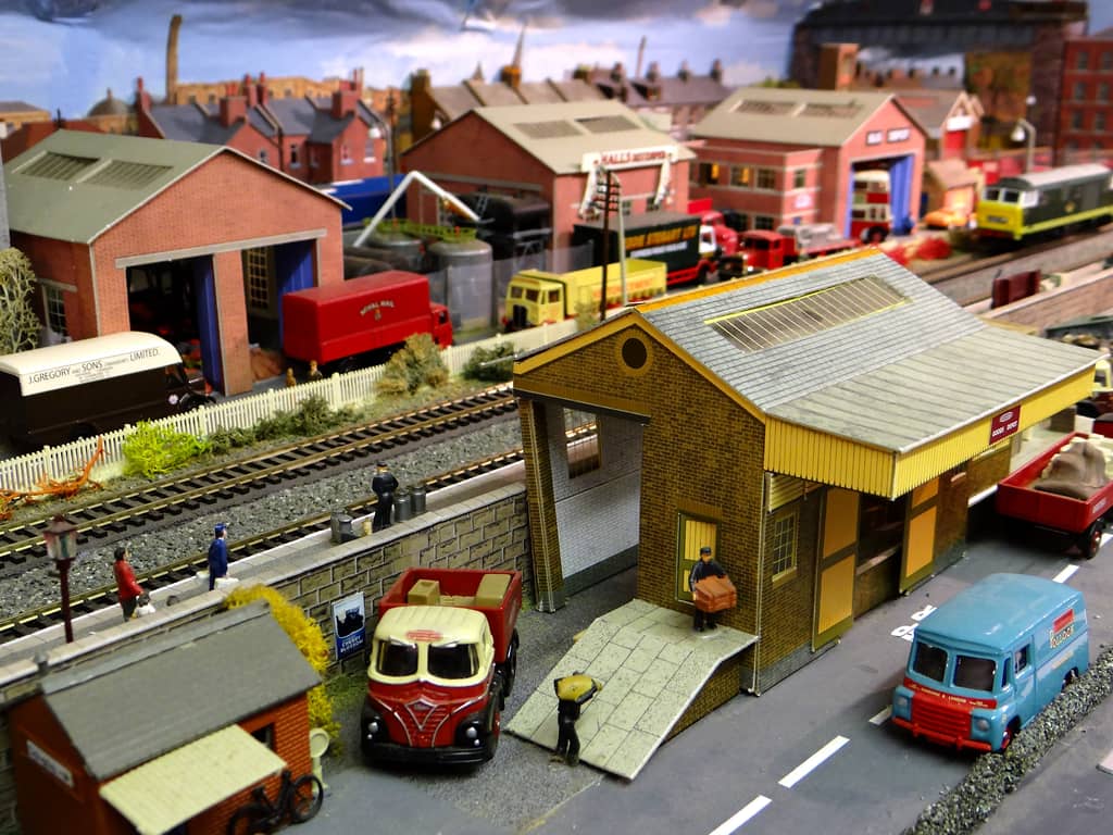 A yellow-brick goods shed with a paved ramp, a porter holding a crate at its door and a man carrying a sack below, a red and cream lorry beside it, and lorries, the bus depot and a green diesel beyond the tracks.