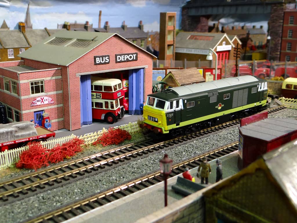 A green diesel locomotive with a yellow front, numbered D7046, stands on the far track beside a red-brick bus depot where a red and cream double-decker waits in the open doors, with passengers on a platform in the foreground.