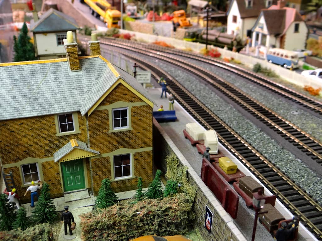 A yellow-brick two-storey house with a green door stands beside a railway platform with luggage barrows, as curving tracks lead past a timber yard and cottages in the distance.