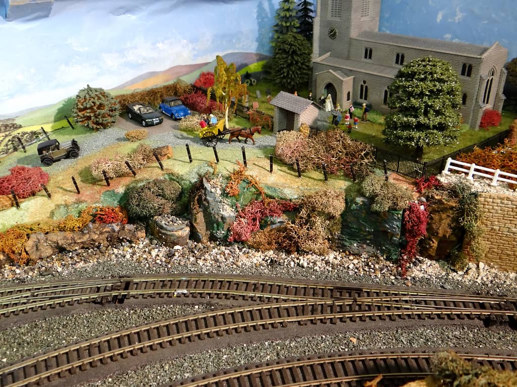 A grey stone church with a clock tower on a grassy hill, with a wedding party at the porch, a pony and trap and parked cars on the lane, above a rocky cutting and railway tracks.