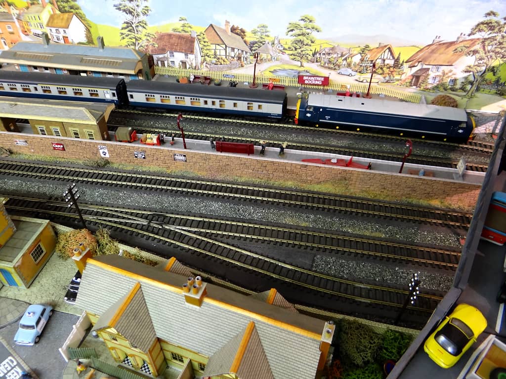 A blue diesel and blue-and-grey coaches stand at a platform with a red nameboard, behind a stone wall with enamel signs, several tracks and the roofs of yellow-brick terraces in front.