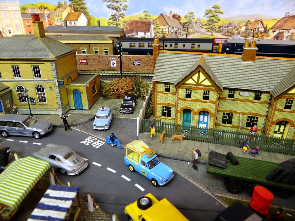 A yellow-brick police station and a row of yellow-brick houses beside a road with two police cars, a light blue ice-cream van and a coal lorry, with a train behind a wall.