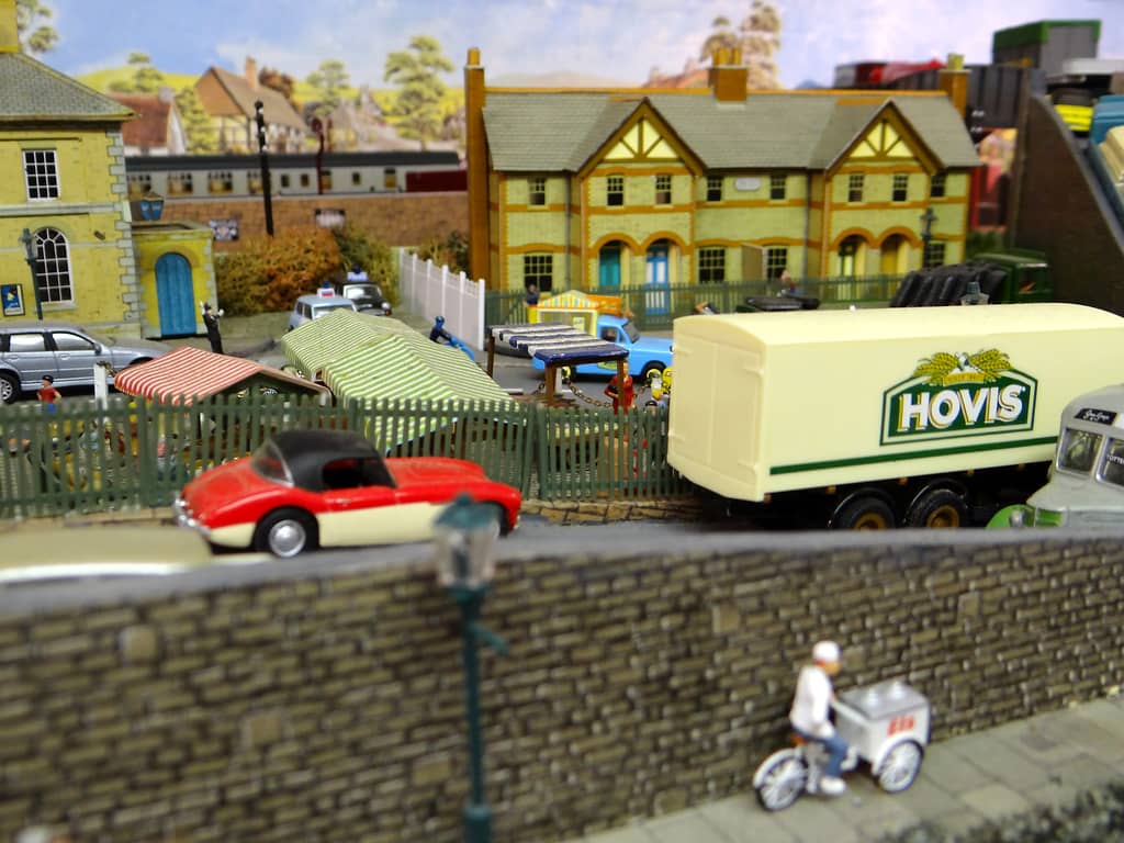 A cream Hovis lorry on a road behind a stone wall, with a red and cream sports car, striped market stalls, and an ice-cream seller on a tricycle on the pavement in front.