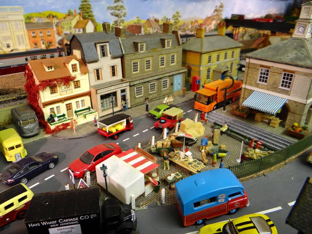 A cobbled market square seen from above, with traders among crates and sacks, a red and white stall, a blue and red circus van and a black lorry, and shops behind.