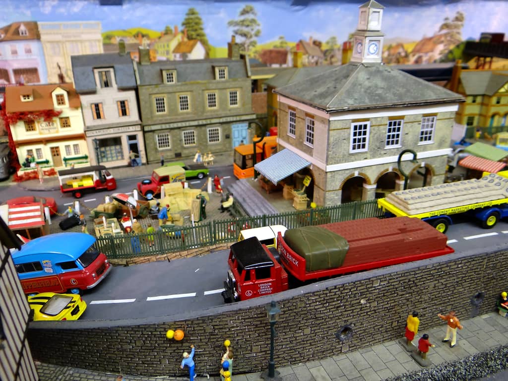 A stone market hall with a clock tower behind a road with a red lorry of bricks and a yellow lorry of cement sacks, with children playing ball on the pavement in front.