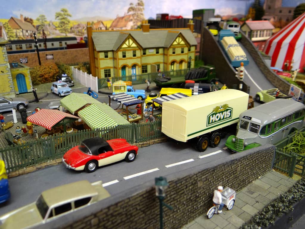 A cream lorry with a green HOVIS sign, a grey and green coach and a red and cream sports car on a road beside striped market stalls, with an ice-cream tricycle in front.