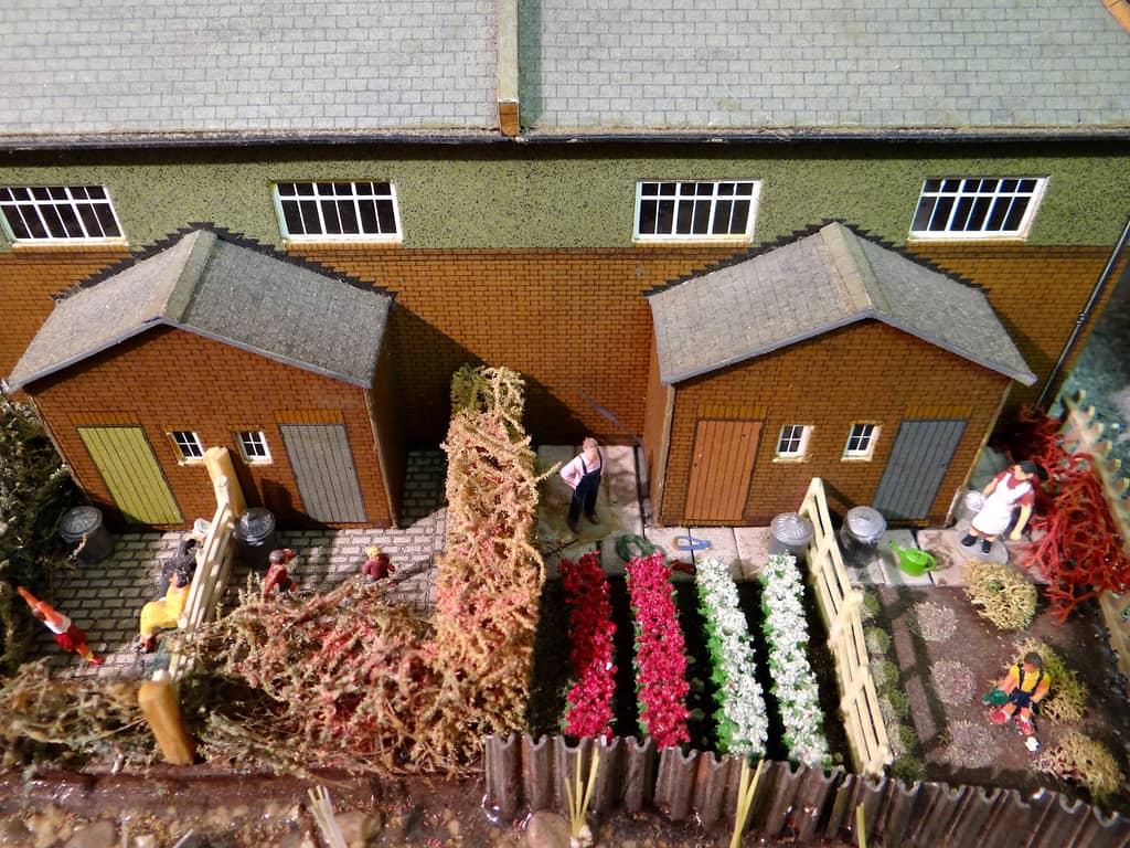 Looking down on the back of a brick terrace with two outhouses, dustbins, rows of red and white flowers and model figures in the yards and garden.